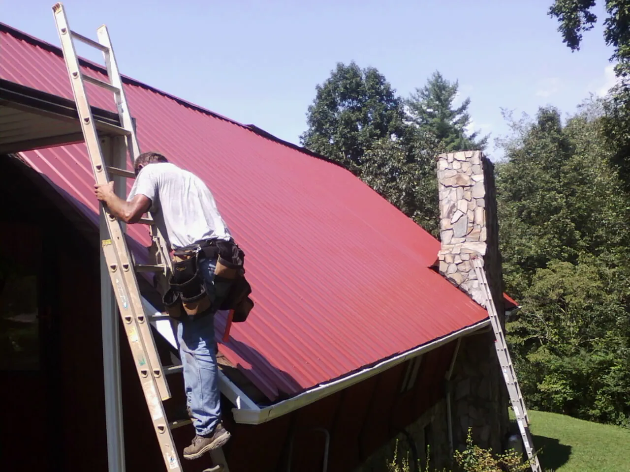 Roofer on a ladder beside a red metal roof and stone chimney — Soffit & Fascia Repair by Roofing Brothers in Mica